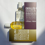Two bottles of MALIN+ Moroccan Fig and Tobacco eau de parfums with their respective purple and gold cardboard boxes, arranged on a white background with sharp shadows.