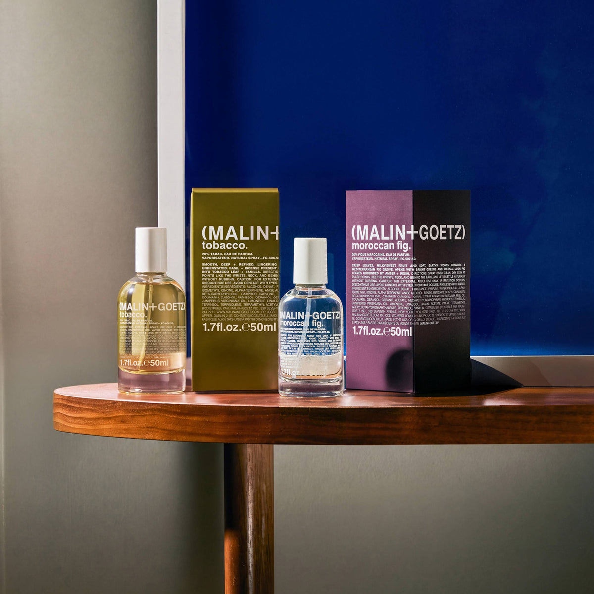 Two MALIN+GOETZ skincare products and their boxes arranged on a wooden table against a split gray and dark blue background. The set includes tobacco and fig eau de parfums.